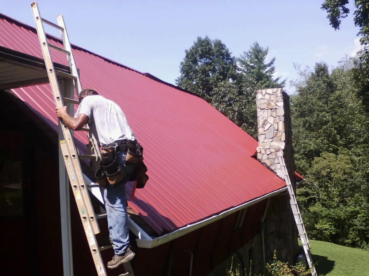Roofer on a ladder beside a red metal roof and stone chimney — Soffit & Fascia Repair by Roofing Brothers in Blue Springs